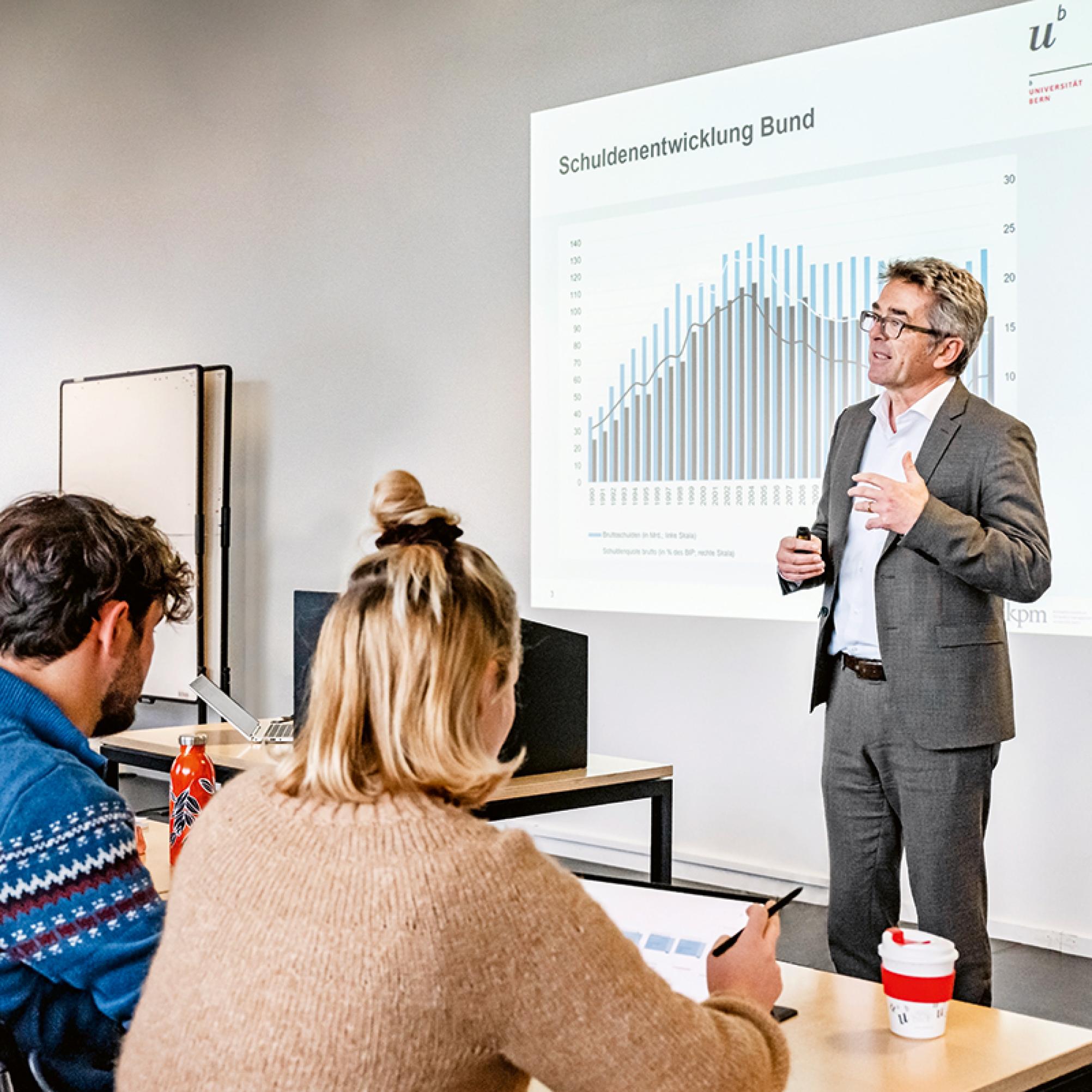 Ein Lehrer zeigt eine Präsentation zur Entwicklung der Schuldenbremse. Studentinnen und Studenten hören ihm zu.
