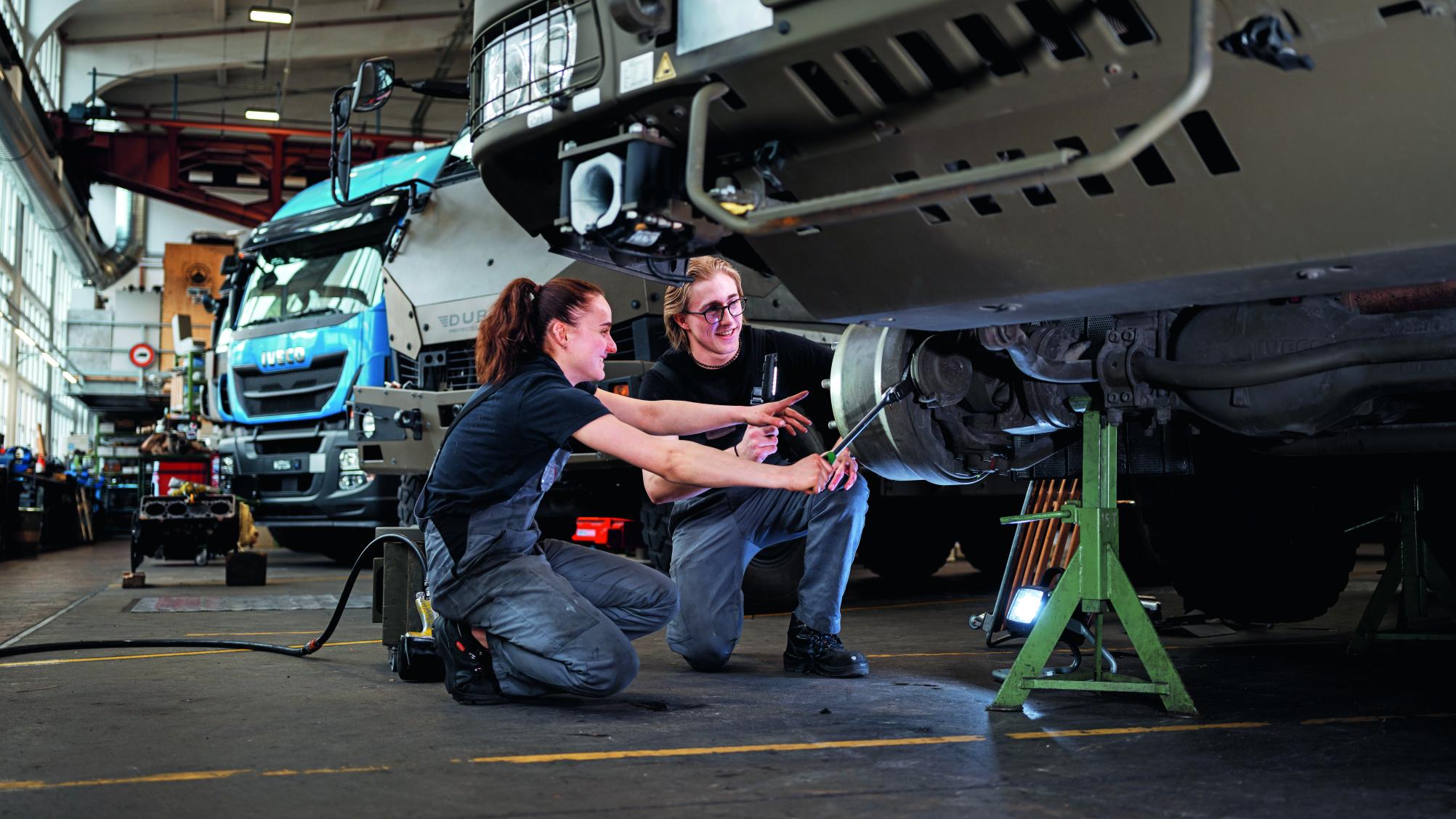 Eine junge Frau in Arbeitskleidung zeigt ihrem Kollegen, wie man das Rad eines Militärfahrzeugs wechselt.